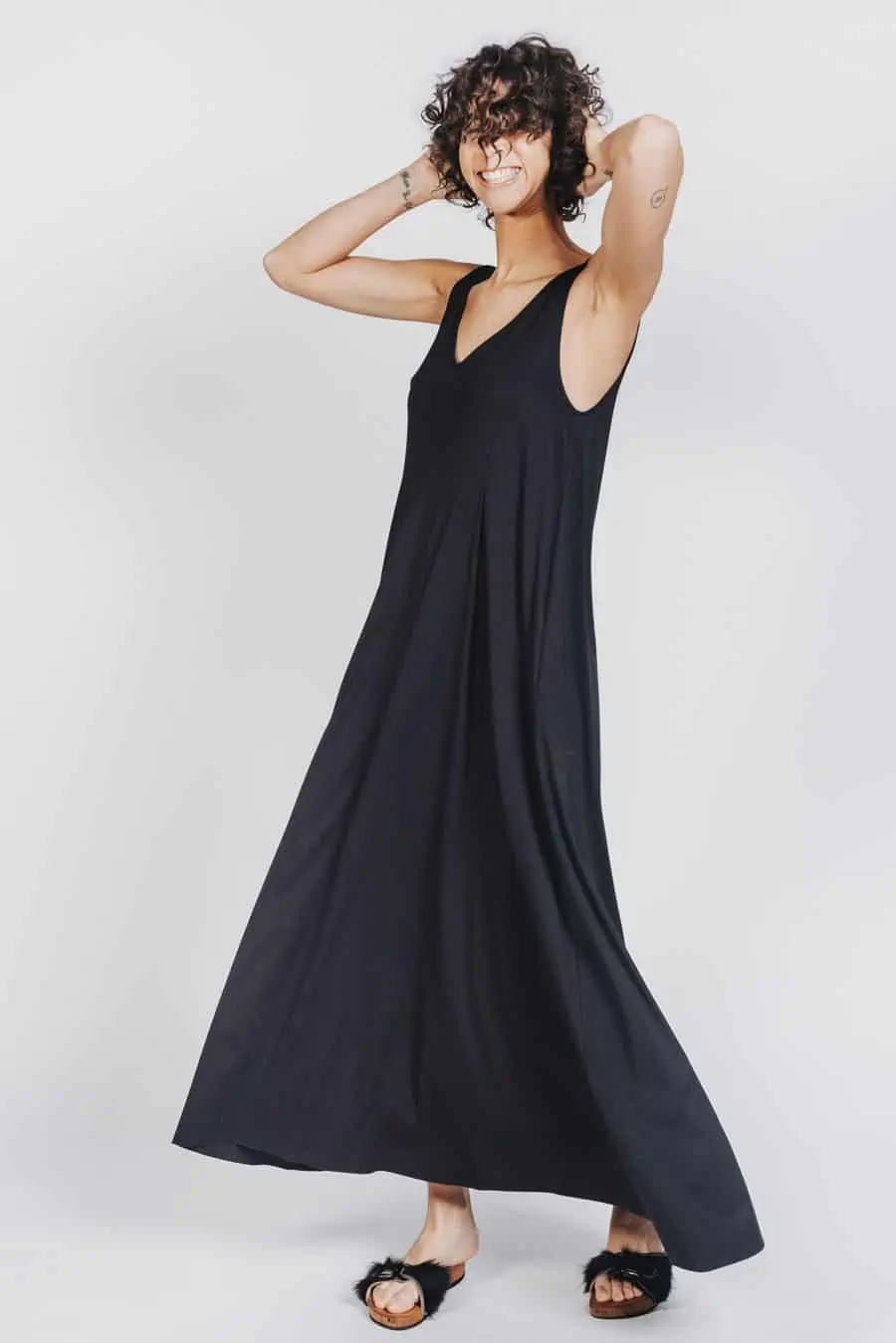 Deep White Black Modell SASHA langes Kleid schwarz mit schwarzen Fellsandalen aus Holz von Devich, ärmelloses Sommerkleid mit V-Ausschnitt, Ansicht gesamt seitlich von vorne